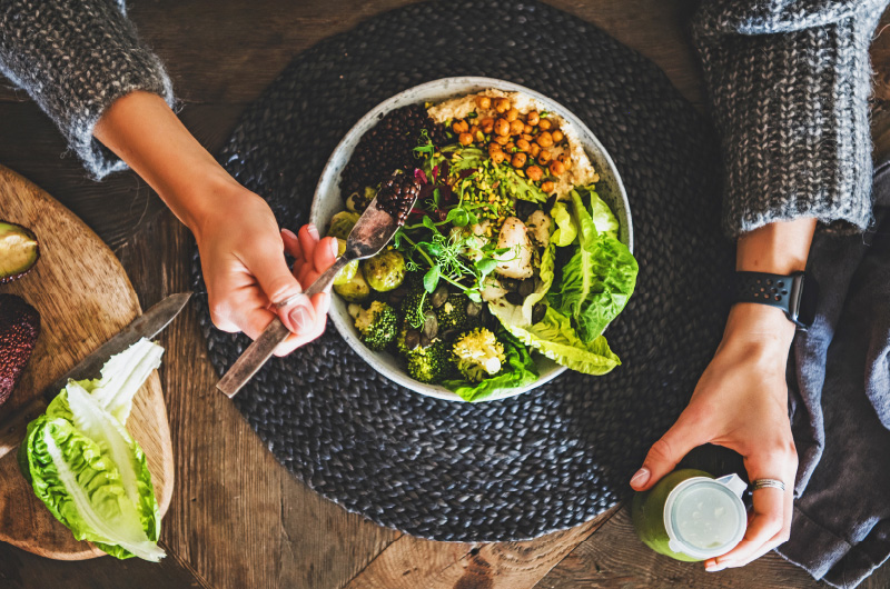 Overhead shot of woman adding blackberries to a green salad with right hand and holding bottle of green dressing in left hand