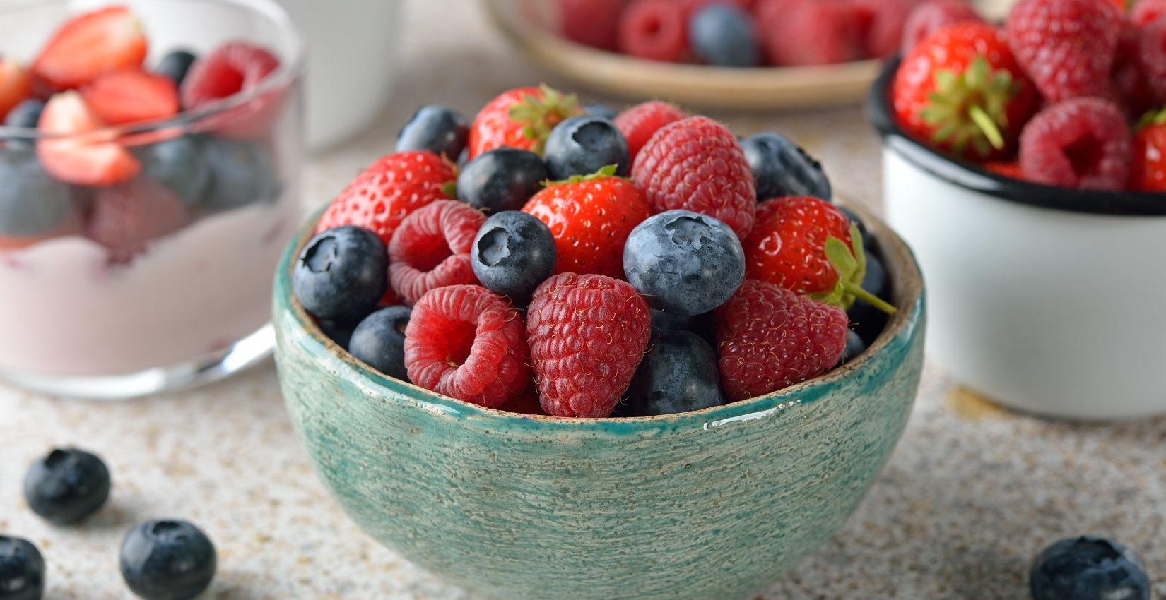 image of strawberry, blackberries, raspberries, and blueberries in a teal ceramic bowl with more in the background